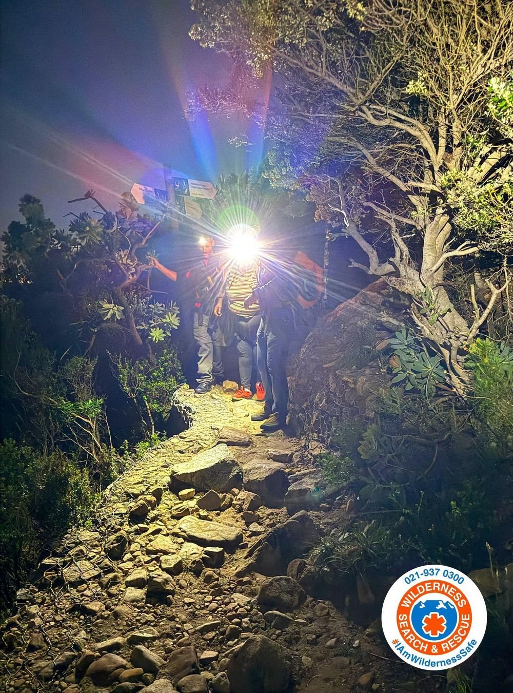 Rescuers with headlamps help a hiker down a rocky, brush-lined trail at night, with the Wilderness Search and Rescue logo in the corner