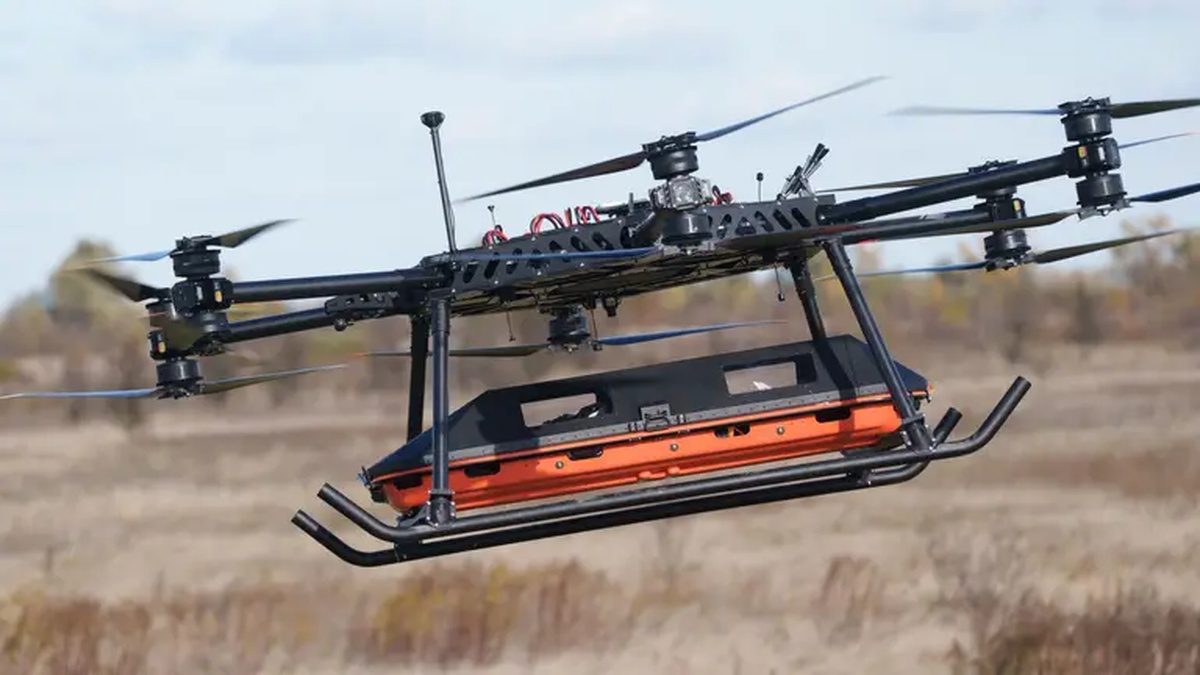 SkyFall’s StarGate, a large multirotor drone with an enclosed orange stretcher compartment slung beneath, flying low over a field