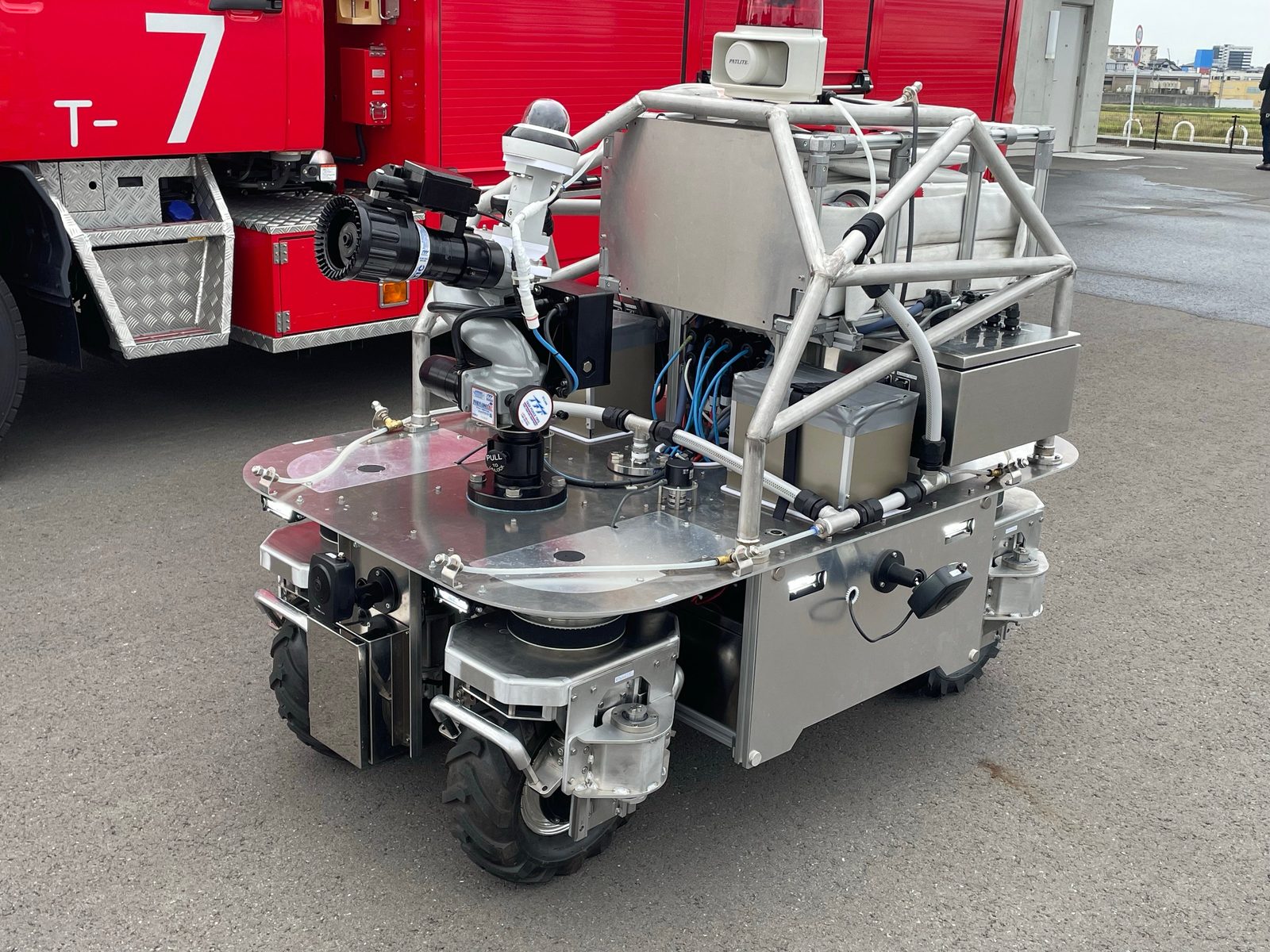 tmsuk’s prototype unmanned water-discharge robot: a four-wheeled stainless-steel chassis with a black water-cannon nozzle, cameras, a hose box and a roll-cage frame, parked beside a red Munakata District Fire Department truck