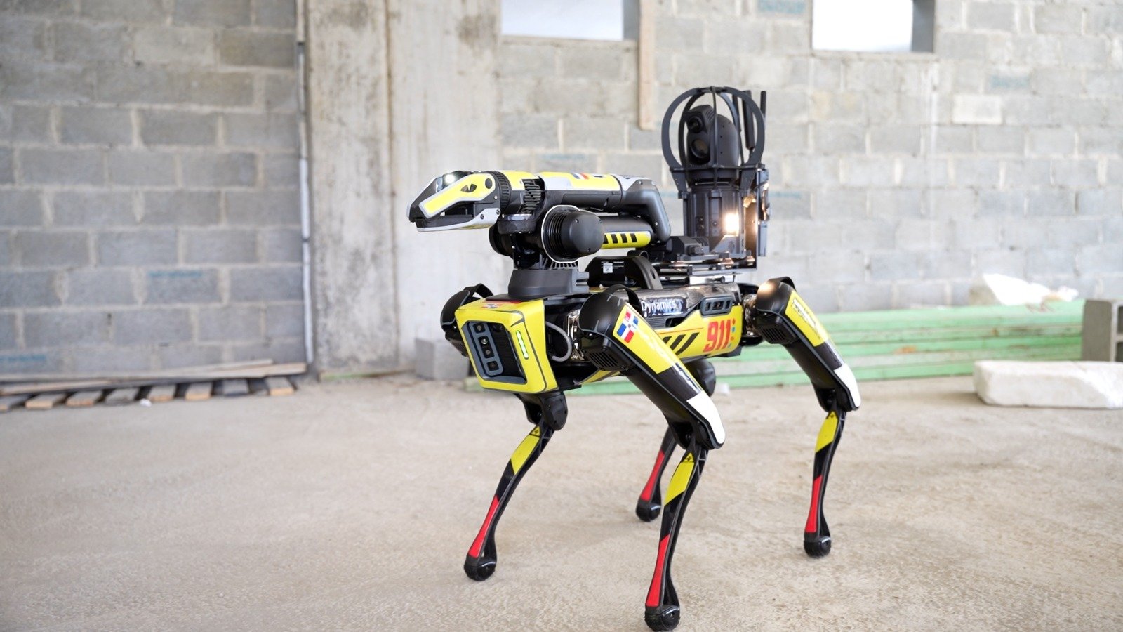 A yellow and black four-legged robot with a mounted camera and sensor head stands on a concrete floor in front of a block wall